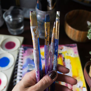 hand holding small paint brushes with paint on them, paint and a canvas are in the background
